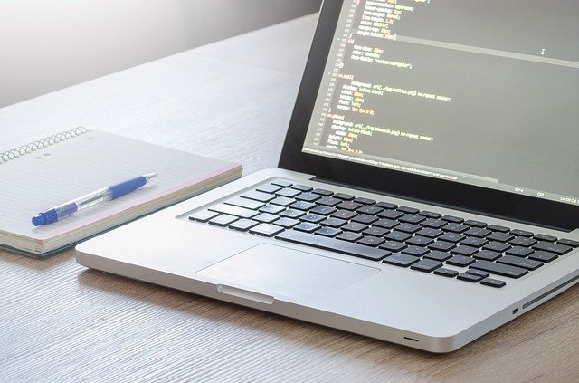 A laptop is on a desk, beside a notepad and pen.  The laptop screen shows coding in process.