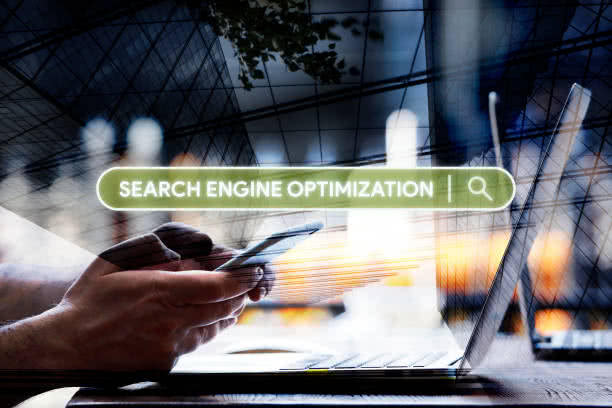 A blurred background and side view of two hands holding a cell phone above a laptop.  Ready to apply Search Engine Optimization to a website.